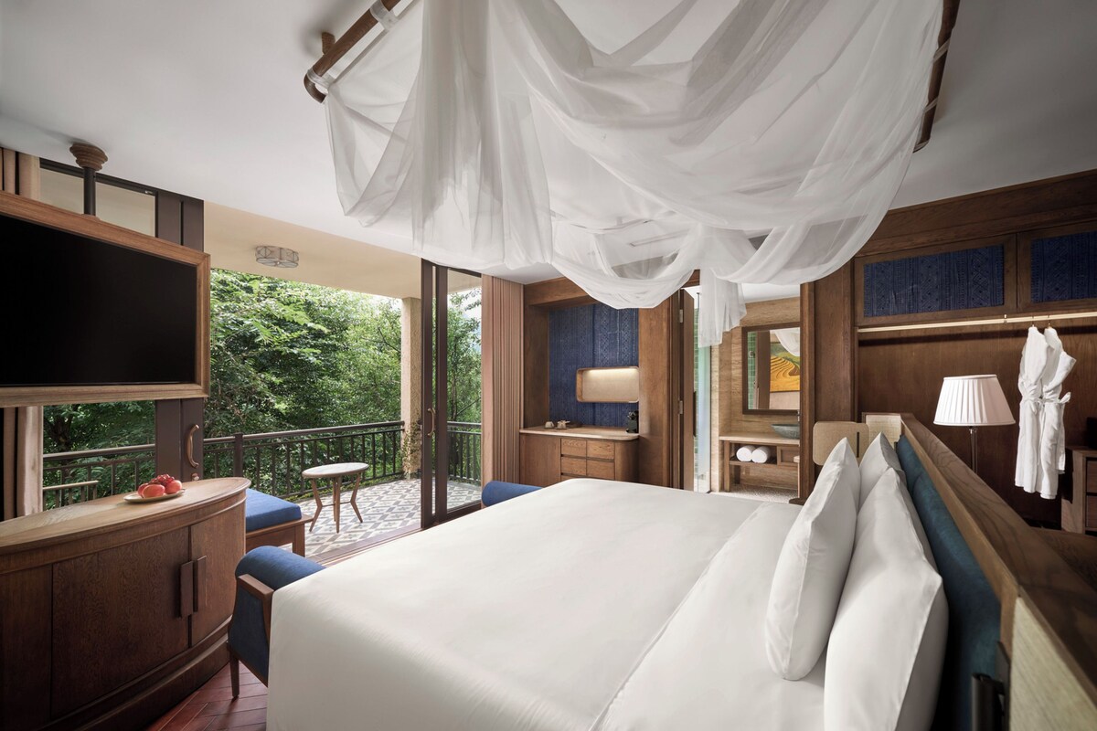 A hotel room with a large bed, a TV, a balcony with a view of greenery, and a wooden desk with a chair.
