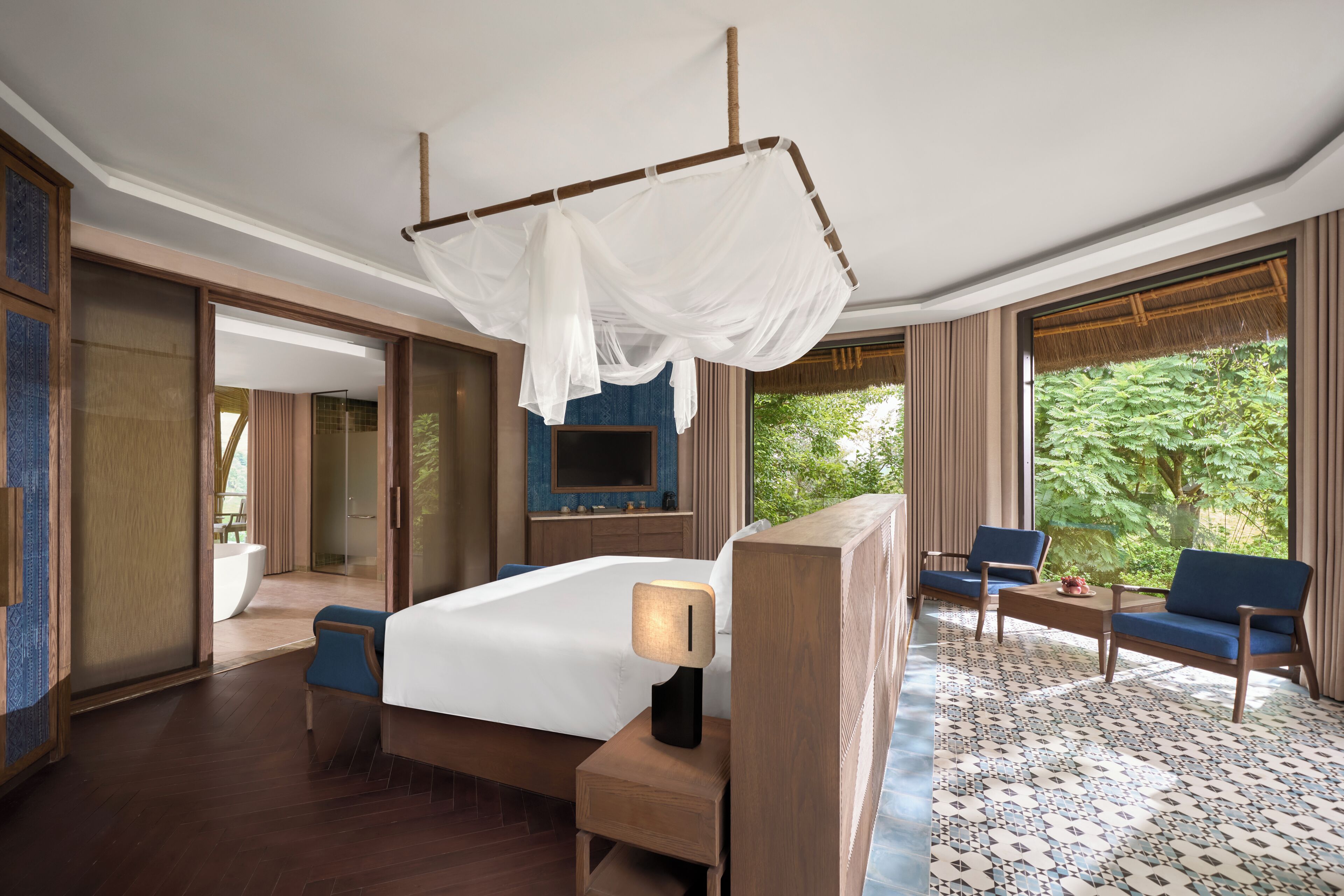 A hotel room with a bed, bedside table, TV, seating area, and large windows.