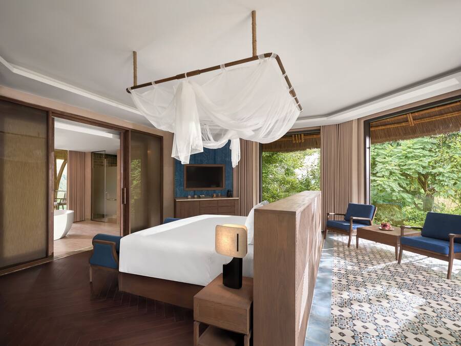 A hotel room with a bed, bedside table, TV, seating area, and large windows.