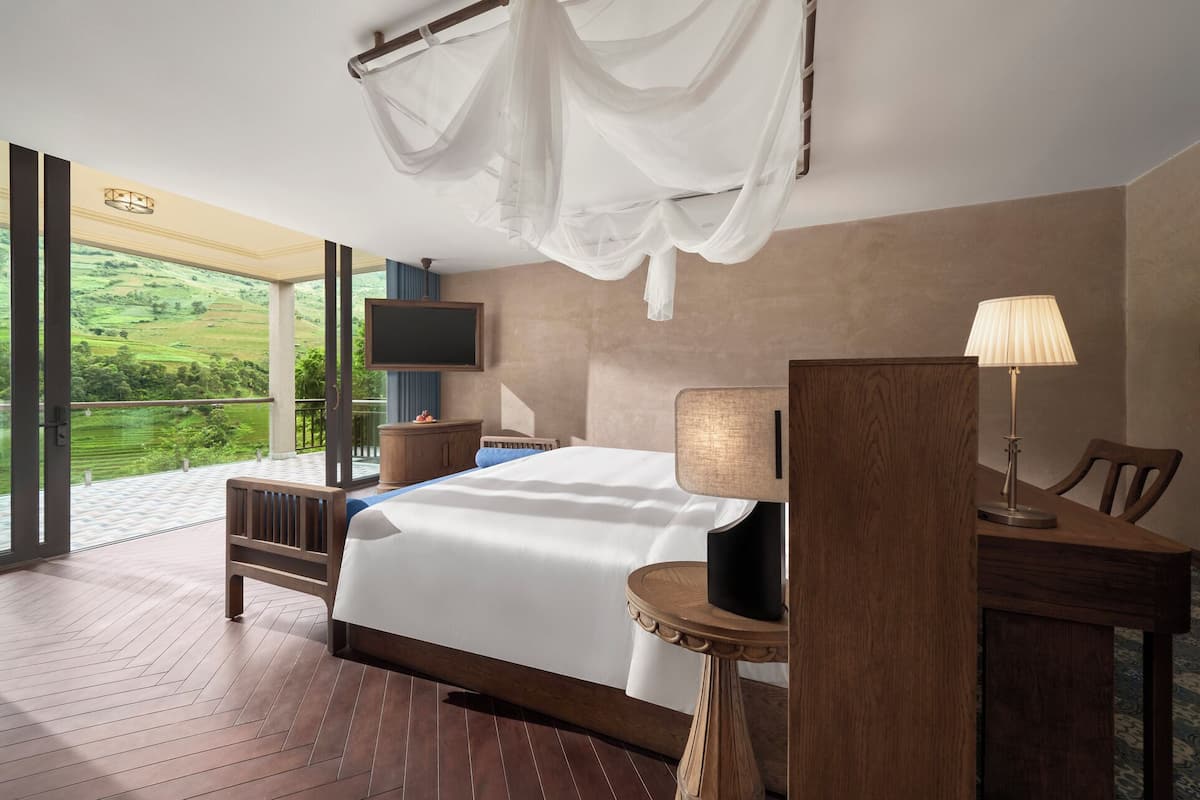 A bedroom with a bed, a desk, a chair, a TV, and a balcony with a view of greenery.