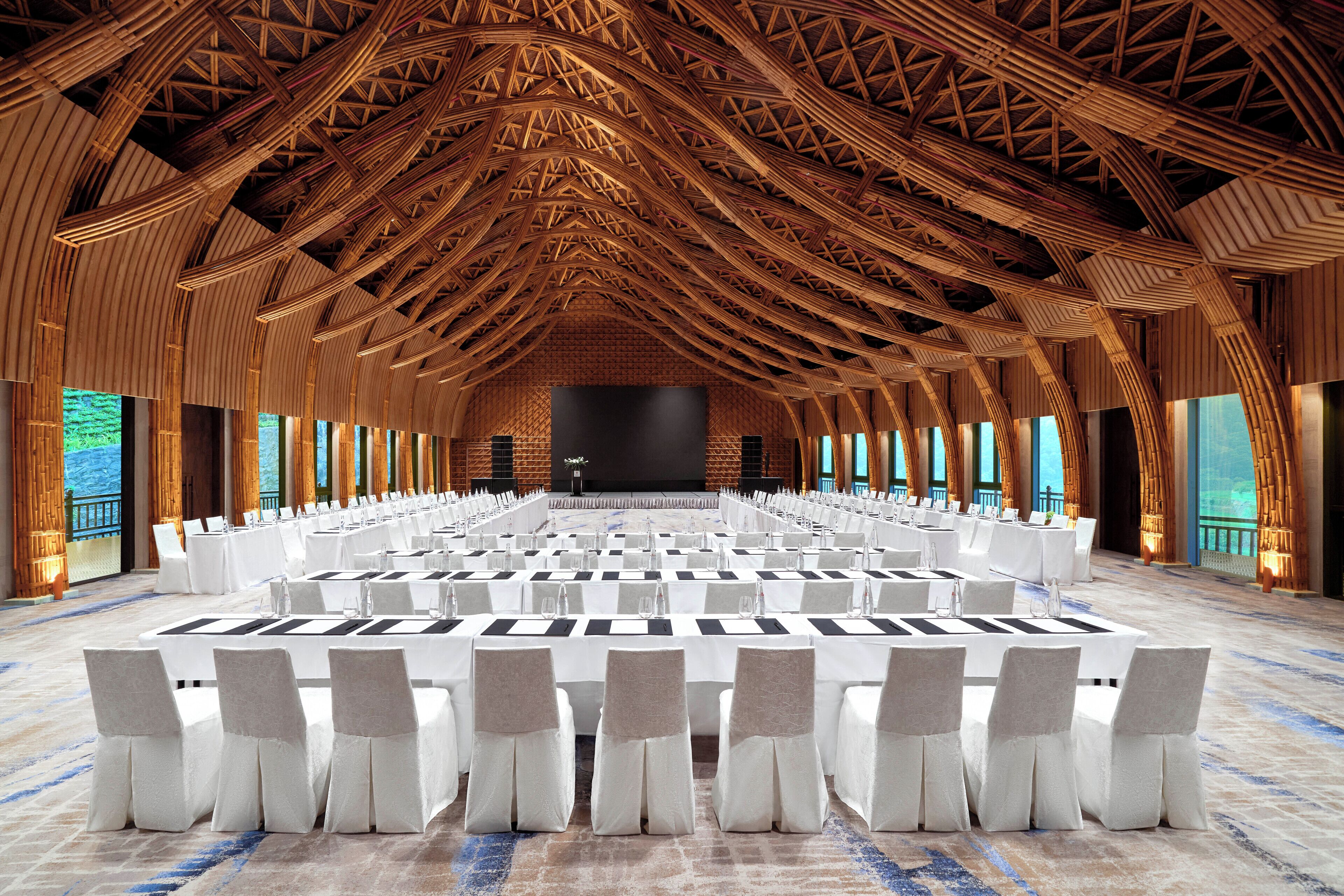 Un amplio salón de conferencias con un techo alto de madera y un escenario en la parte delantera. Las mesas están dispuestas con manteles blancos y sillas.
