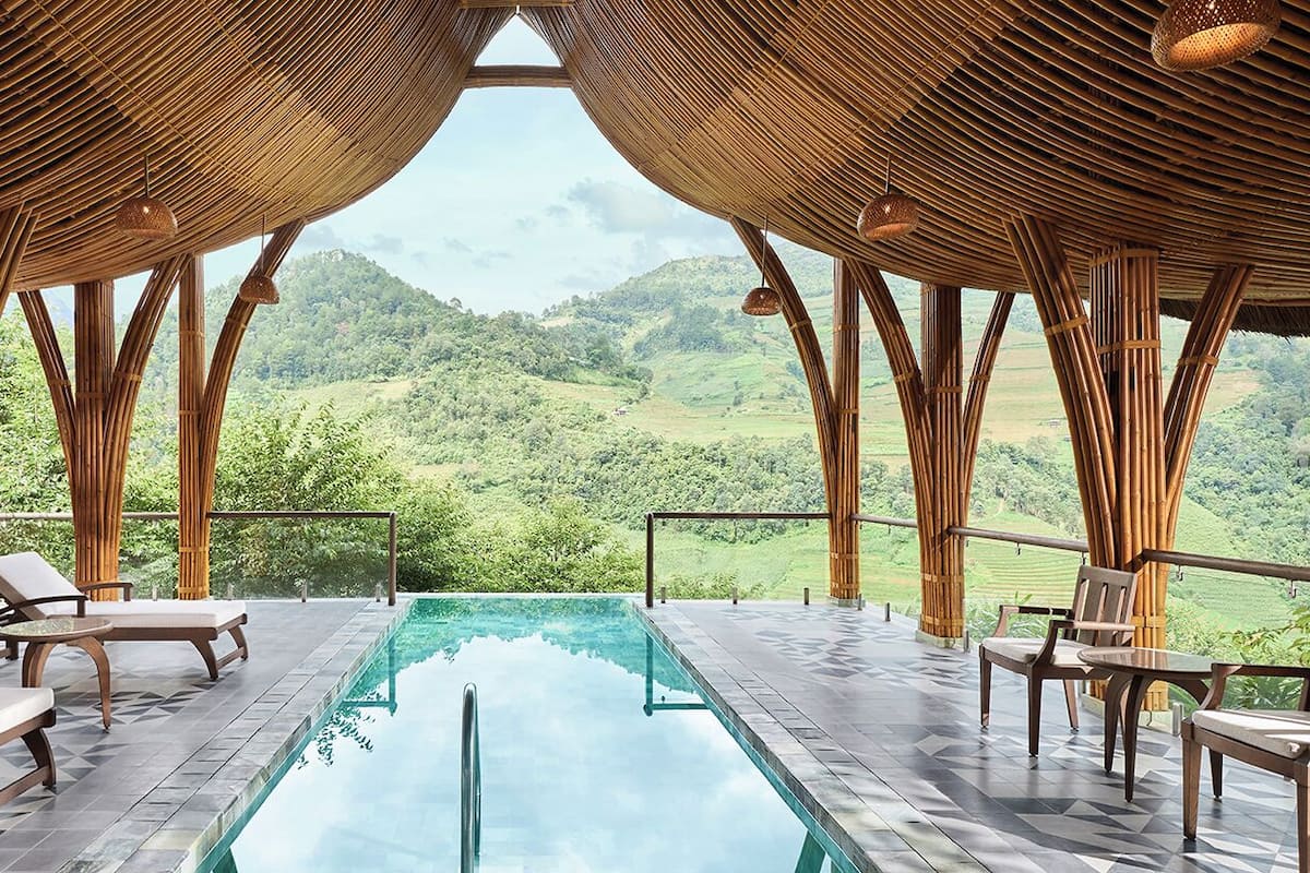 A pool area with lounge chairs and tables, surrounded by bamboo structures, overlooking a lush green landscape.