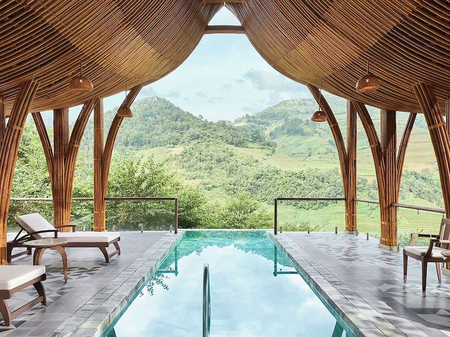 A pool area with lounge chairs and tables, surrounded by bamboo structures, overlooking a lush green landscape.