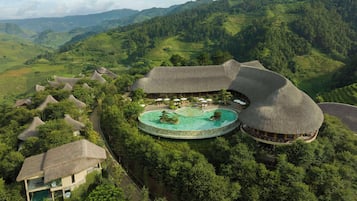 A resort with thatched-roof villas, a central pool, and lush green hills in the background.