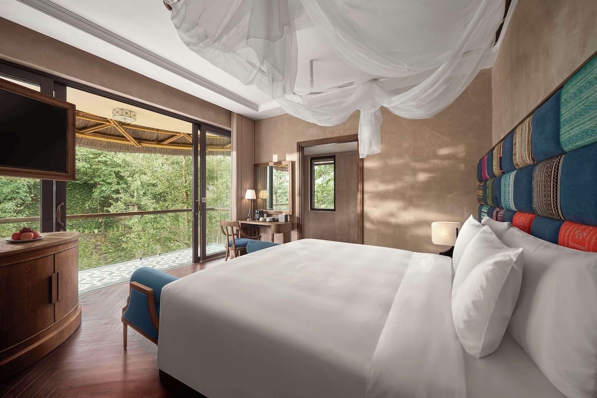 A bedroom with a large bed, a desk, and a TV. There is a balcony with a view of trees.
