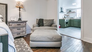 Un salon confortable avec un canapé, une lampe et une table d’appoint. On aperçoit une cuisine à travers une porte ouverte.