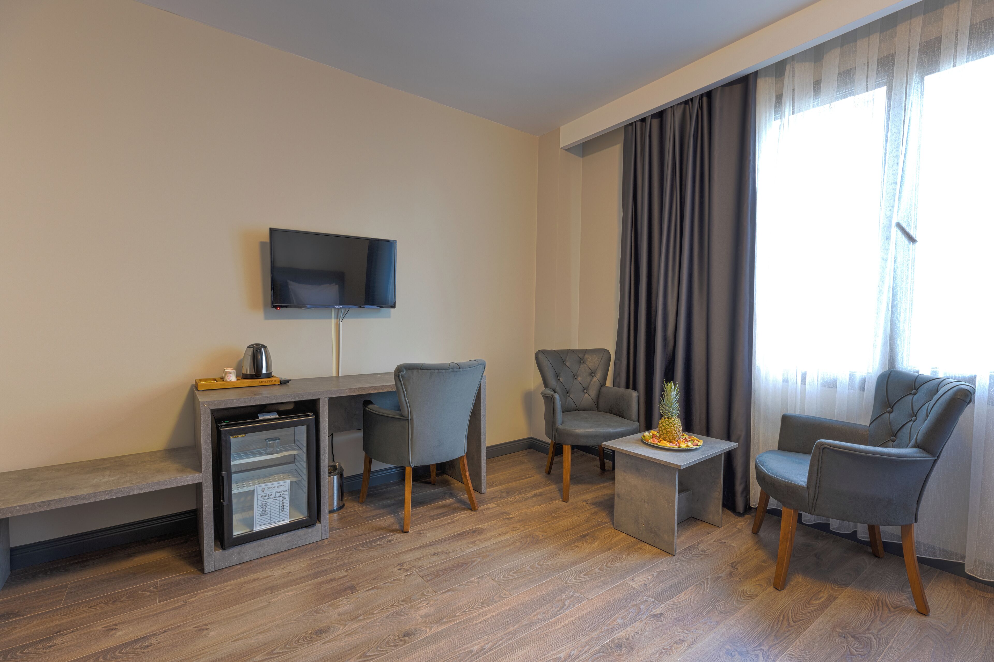 A modern hotel room with a flat-screen TV mounted on the wall, a small refrigerator, a wooden coffee table, two grey armchairs, and a sofa.