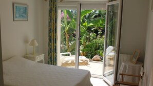 Une chambre à coucher comprenant un lit, une table de chevet, une lampe et une porte-fenêtre coulissante donnant sur une terrasse avec une chaise et des plantes.