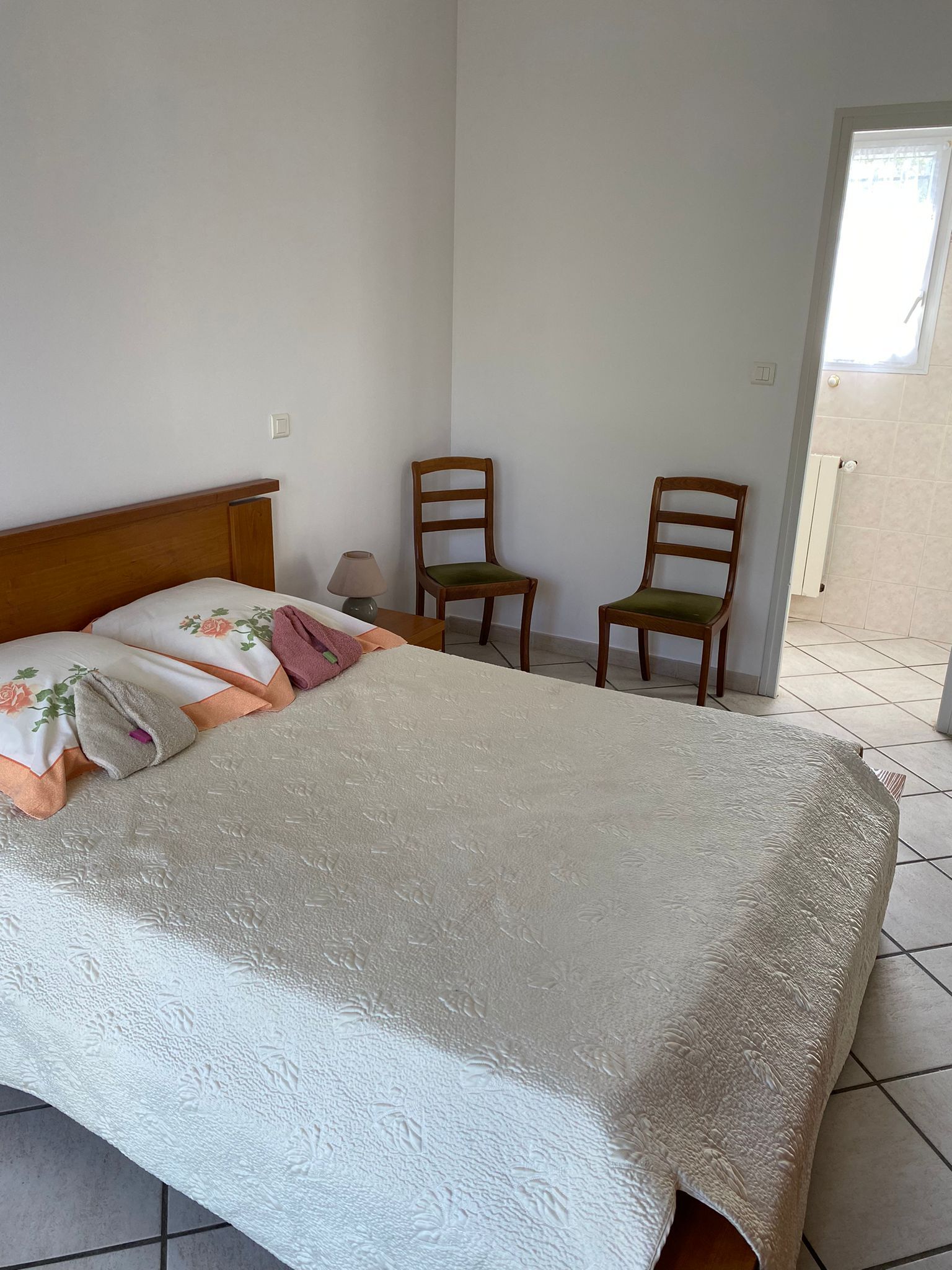 A bedroom with a bed, two chairs, a lamp, and a window.