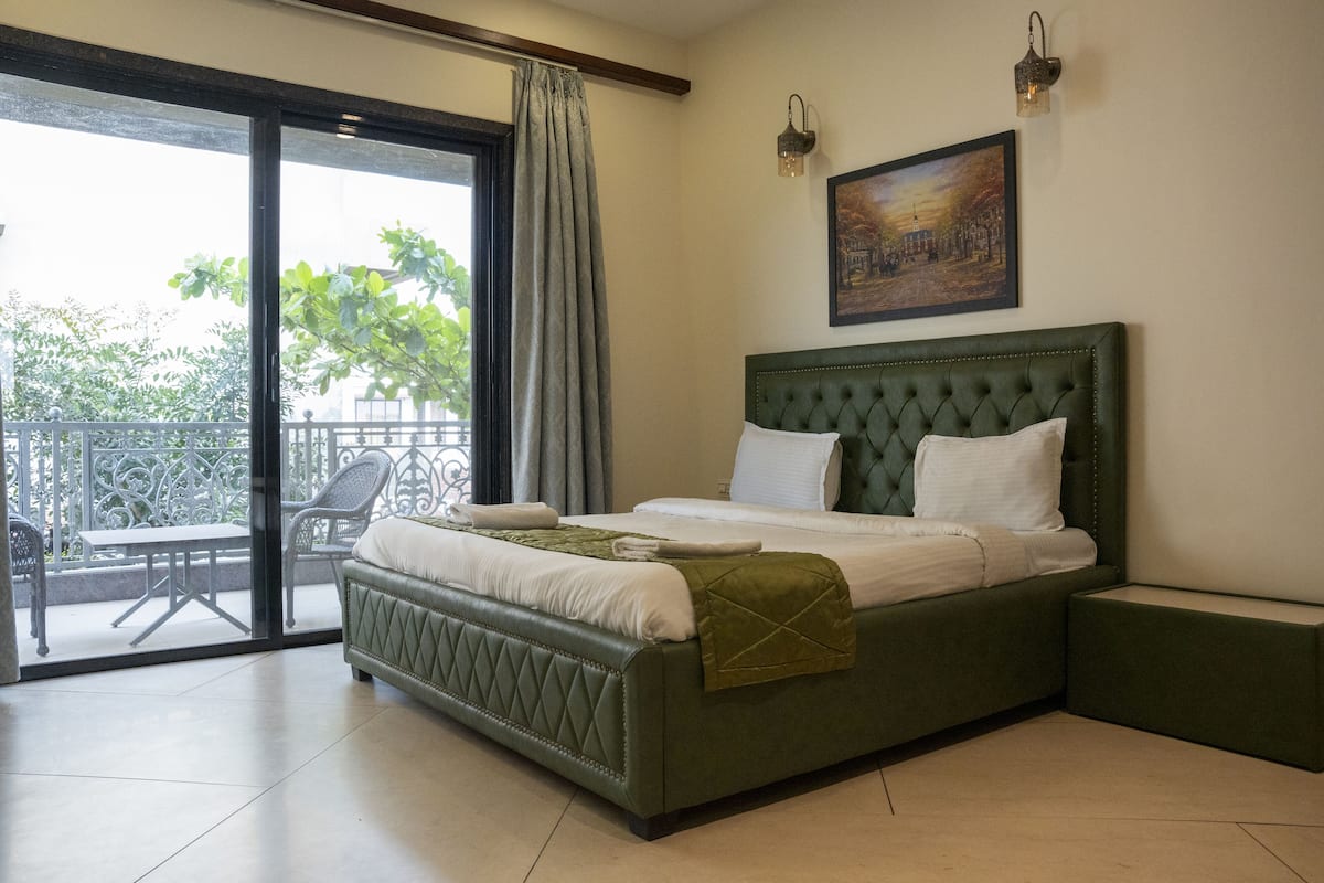 Une chambre à coucher avec un lit recouvert d’un tissu vert, un balcon avec une table et des chaises, et un tableau accroché au mur.