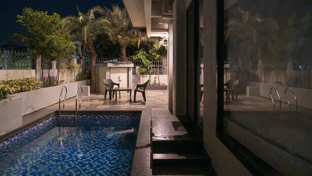 Ein Schwimmbecken mit Fliesenboden, neben einer überdachten Sitzgelegenheit mit Stühlen und Tisch, umgeben von Pflanzen und einem Zaun.