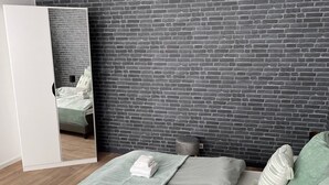 A modern bedroom with a brick-patterned wall, a bed with a teal bedspread and white pillows, a bedside table, and a mirror reflecting the room. - Modernes, Großes City Apartment 95 qm in Meuselwitz (Meuselwitz)