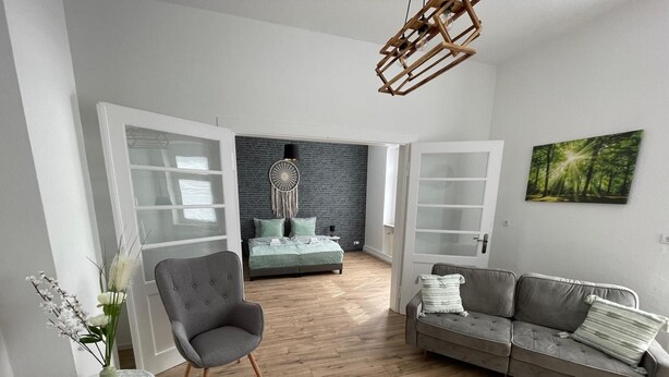 A modern living room with a grey sofa, a grey armchair, a small side table with a vase, and a bed with a grey headboard in the background. - Modernes, Großes City Apartment 95 qm in Meuselwitz (Meuselwitz)