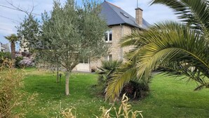A stone house with a grey roof, surrounded by a well-maintained lawn and various trees, including a palm tree. - Appartement à 80 Mètres de la Mer, Chez L'habitant, en Rez-de-jardin (Saint-Lunaire)