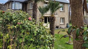 A two-story stone house with a grey tiled roof, surrounded by lush greenery and flowering plants. - Appartement à 80 Mètres de la Mer, Chez L'habitant, en Rez-de-jardin (Saint-Lunaire)