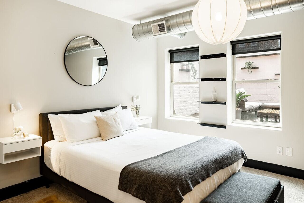 Une chambre moderne avec un grand lit, une table de chevet, un miroir et une fenêtre donnant sur l’extérieur.