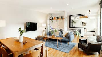 A modern living room with a wooden dining table, a sofa, and a leather armchair. There is a TV mounted on the wall, a plant on the table, and a mirror above the sofa.