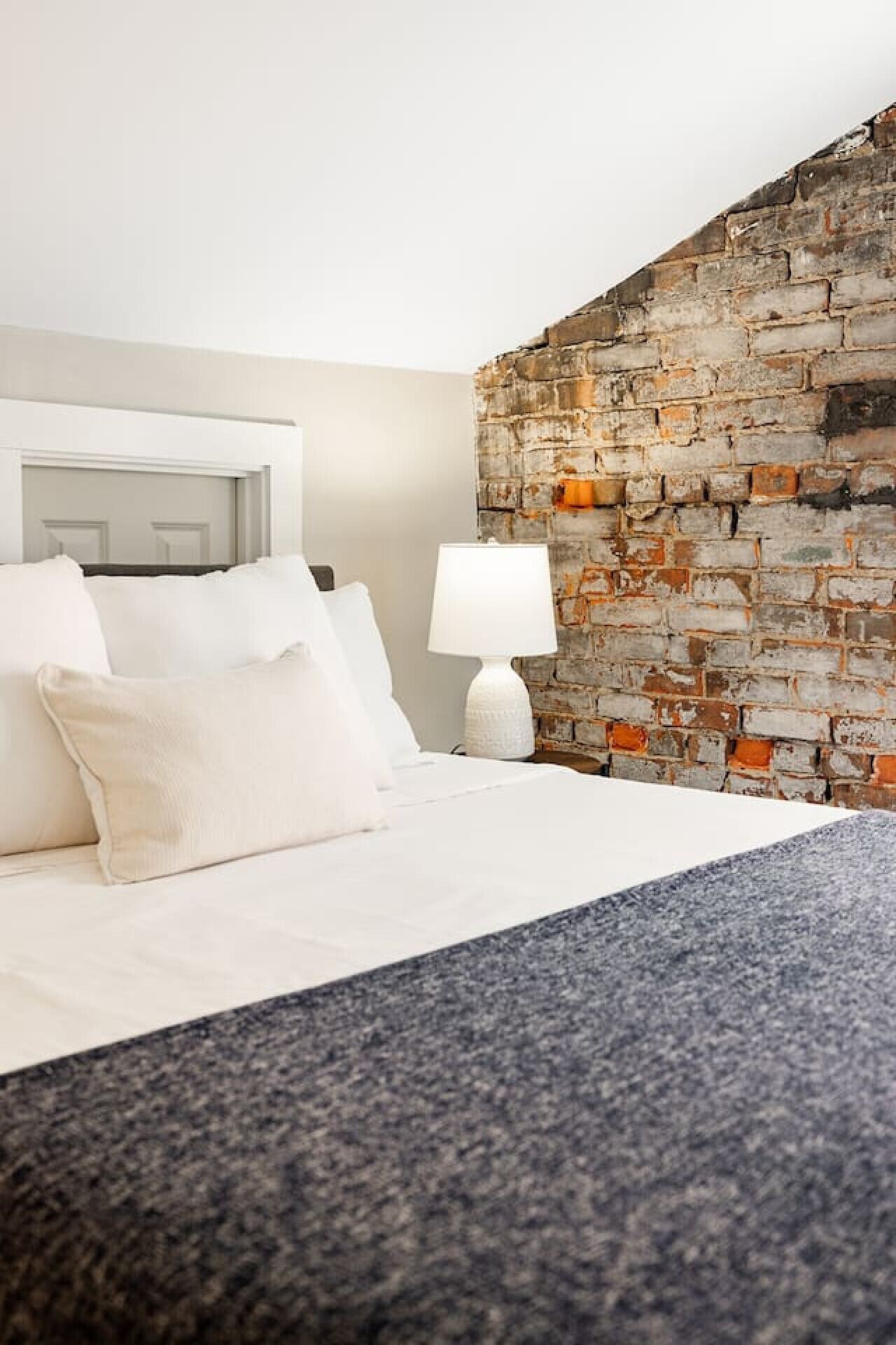 Een hotelkamer met een bed, witte kussens, een witte lamp en een stenen muur.