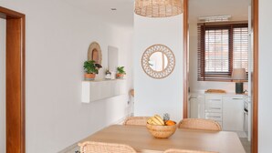 Uma sala de jantar com uma mesa de madeira, cadeiras de vime e uma tigela de frutas. Há plantas em vasos numa estante e uma janela com persianas.