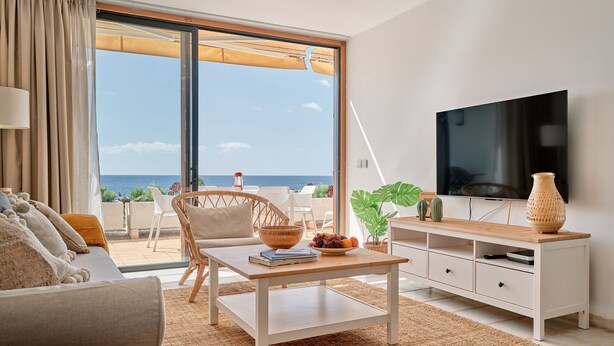 Uma sala de estar moderna com uma televisão grande, uma mesa de centro de madeira e vista para o oceano através de grandes portas de correr.