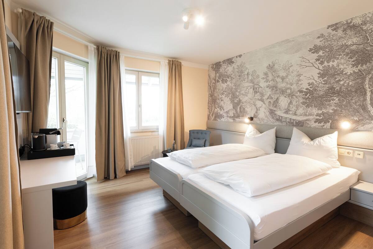 A modern bedroom with a large bed, bedside tables, a nightstand, a desk with a chair, and a window with curtains.