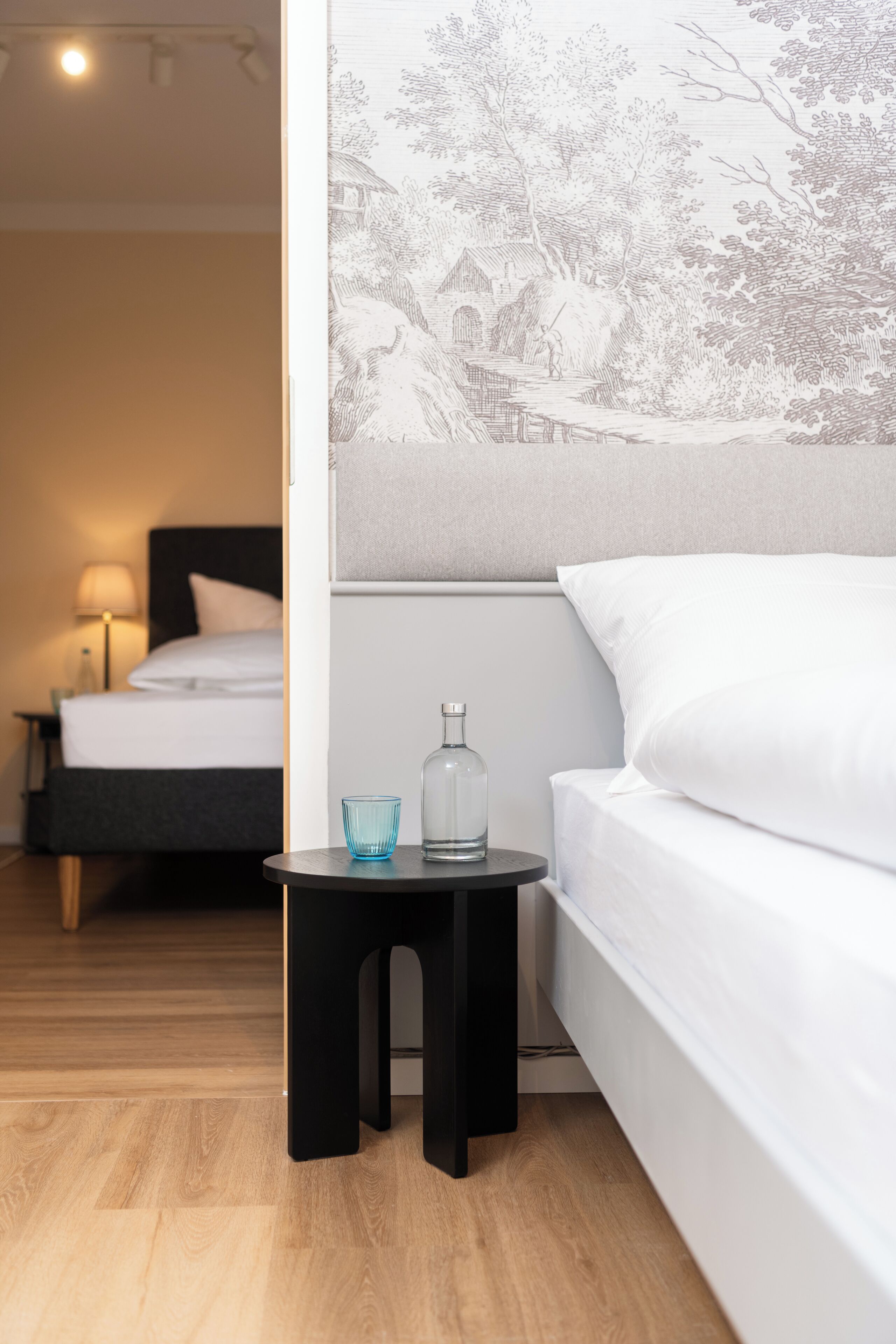 Habitación de hotel con una cama, una mesita de noche con un vaso y una botella, y una lámpara.
