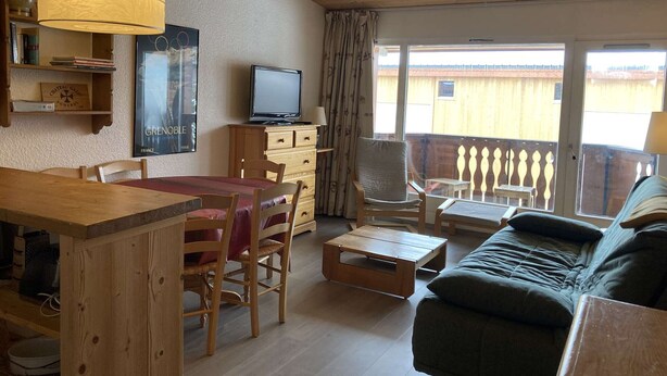 A cozy living room with a wooden ceiling, a dining area with a table and chairs, a sofa, a television, and a balcony with a railing.