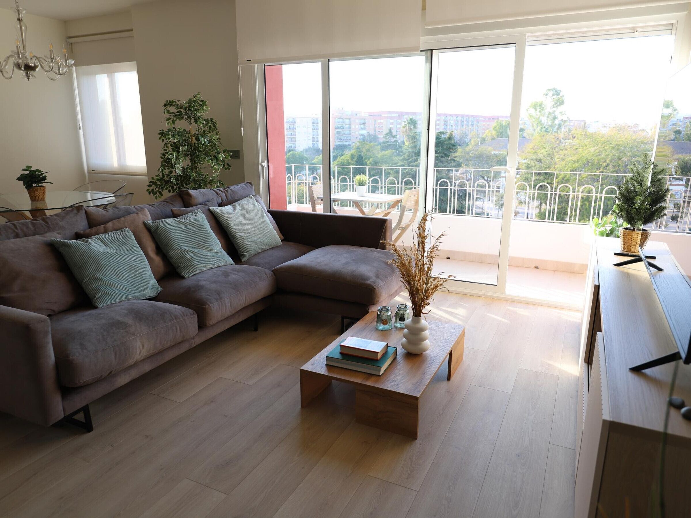 Een moderne woonkamer met een bruine hoekbank, een houten salontafel en een balkon met uitzicht op gebouwen en bomen.