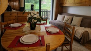 A dining area with a round wooden table set for four, a vase with white flowers, and a view of greenery outside.
