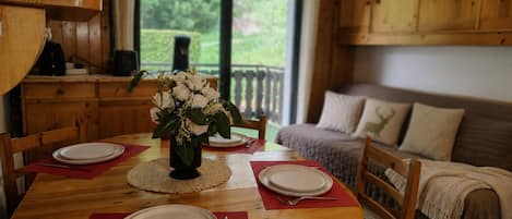 A dining area with a round wooden table set for four, a vase with white flowers, and a view of greenery outside.