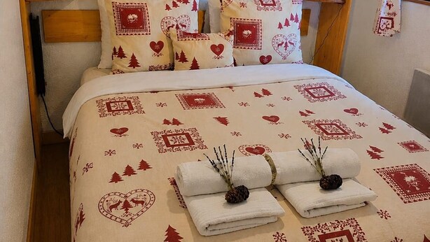 A bed with a patterned bedspread, pillows with heart and tree designs, and two rolled towels with pinecones.