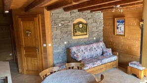 A cozy room with a stone wall, wooden ceiling beams, a sofa with patterned cushions, a small table with a lace tablecloth, and a framed picture on the wall. - Studio with terrace, 4 pers, pets allowed, parking (Châtel)