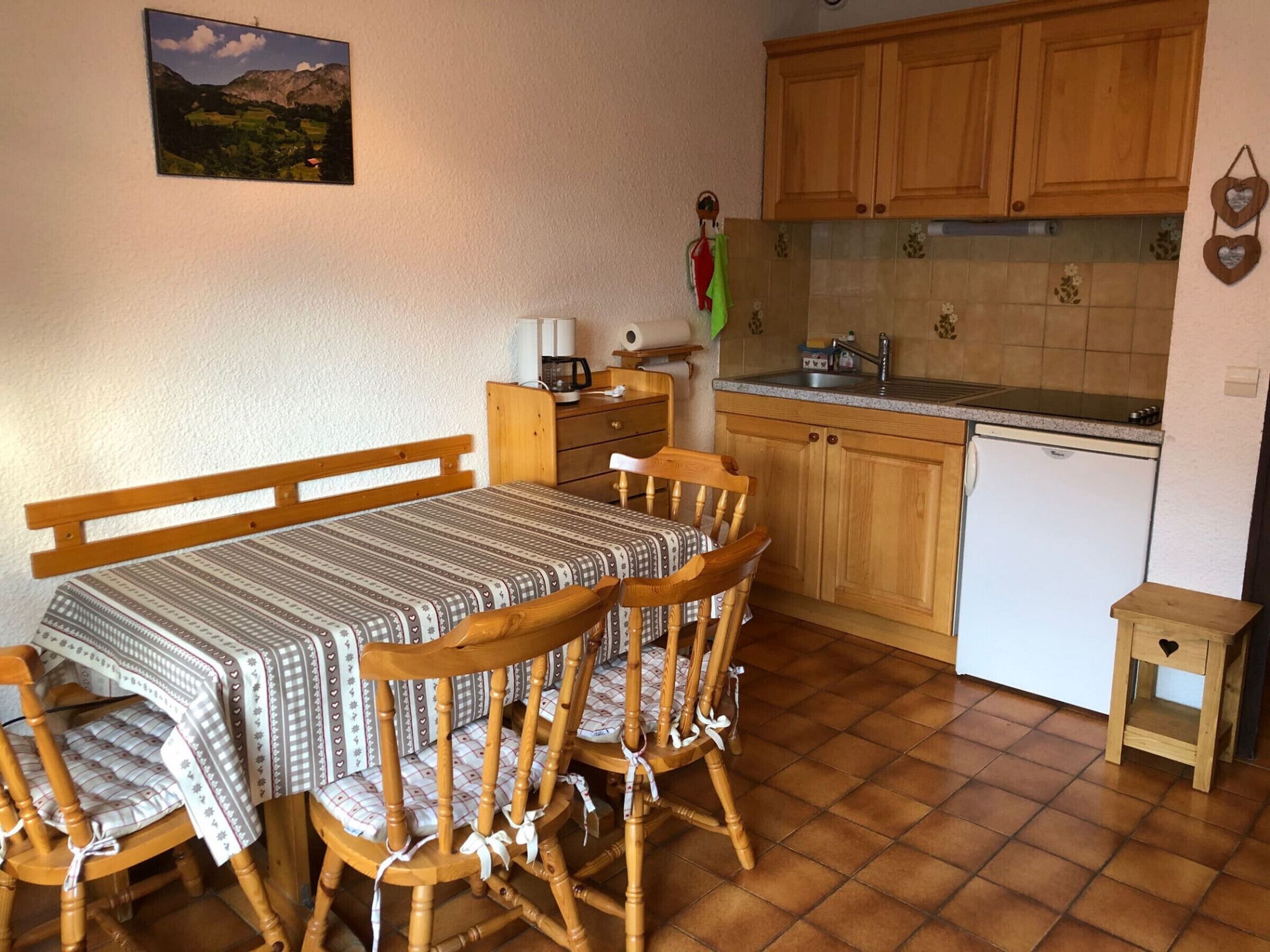 A small kitchen with wooden cabinets, a sink, and a refrigerator. There is a dining table with chairs and a bed with a wooden headboard.
