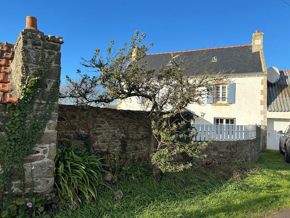 Eine Steinmauer mit Efeu, ein Baum und ein weißer Zaun vor einem Haus mit Ziegeldach und Schornstein.