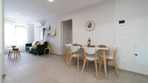A modern living room with a green sofa, a dining table with chairs, and a kitchen area visible through an open door.