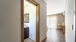 Un interior moderno con un marco de madera en la puerta que conduce a un baño, una zona de comedor con una mesa y sillas, y un gran ventanal con persianas.