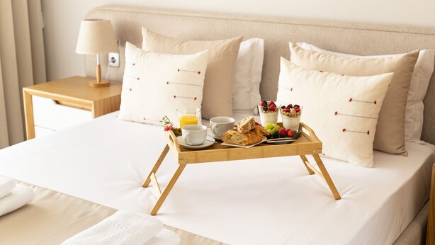 Una cama bien hecha con una bandeja de desayuno, almohadas, una mesita de noche con una lámpara y una ventana con cortinas.