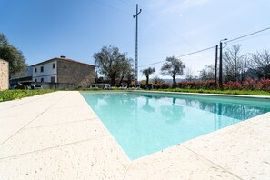 Piscina com deck revestido, rodeada por vegetação e um edifício de pedra.