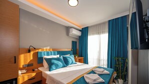 A modern hotel room with a wooden headboard, a bed with blue bedding, a bedside table with a phone and lamp, and a window with curtains. - Otel Carpediem Gold Kuşadası (Kusadasi)