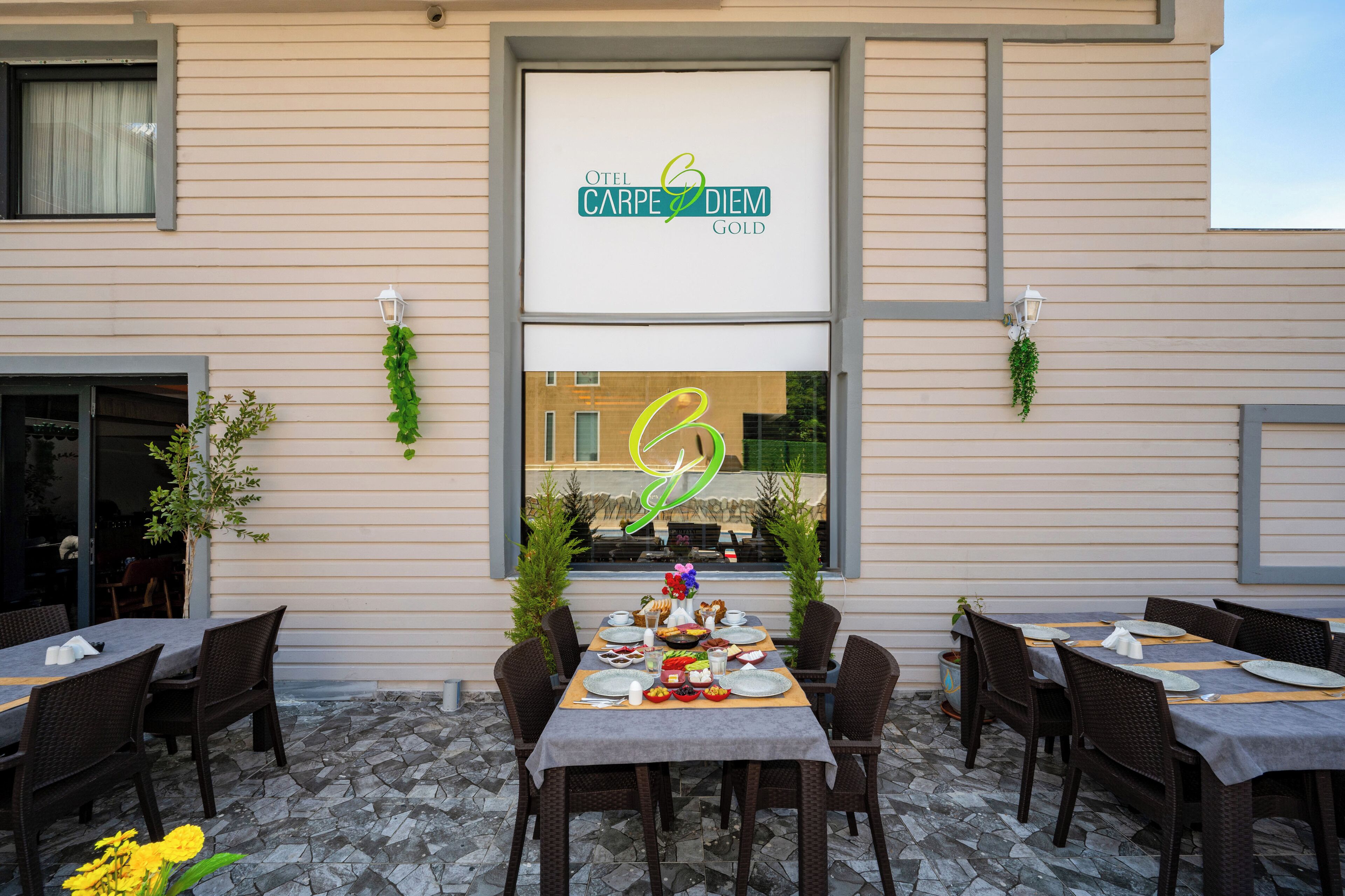 An outdoor dining area with a table set for a meal, surrounded by wicker chairs, and a sign above reading "OTEL CARPE DIEM GOLD".