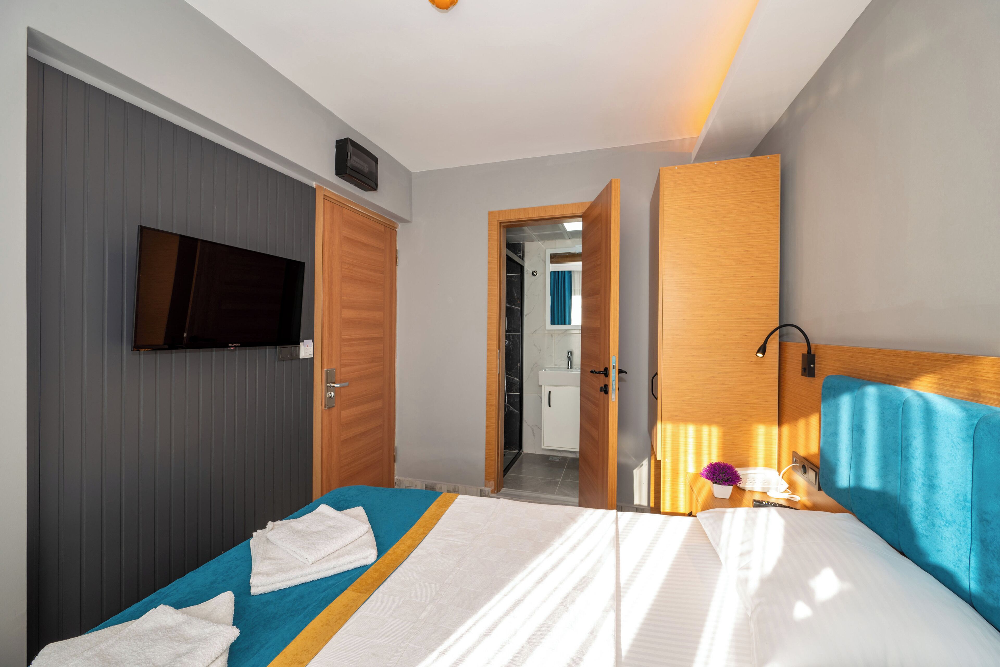 Habitación de hotel con una cama, un televisor montado en la pared, cabecera de madera y un baño visible a través de una puerta abierta.