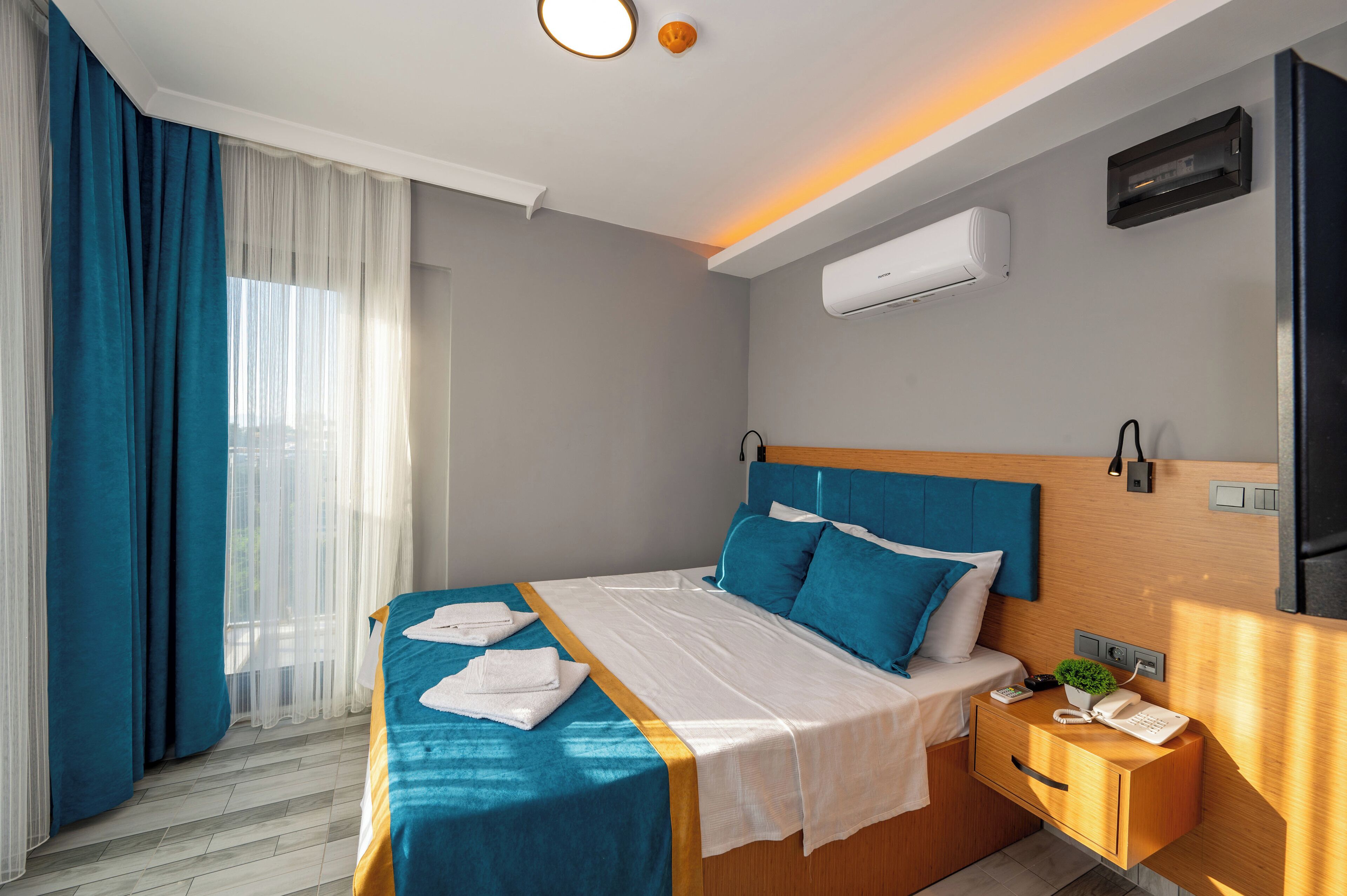 A hotel room with a bed, a nightstand, an air conditioning unit, and a window with curtains.