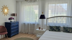 A bedroom with a bed, a dresser, a chair, a nightstand, a lamp, and a window with sheer curtains.