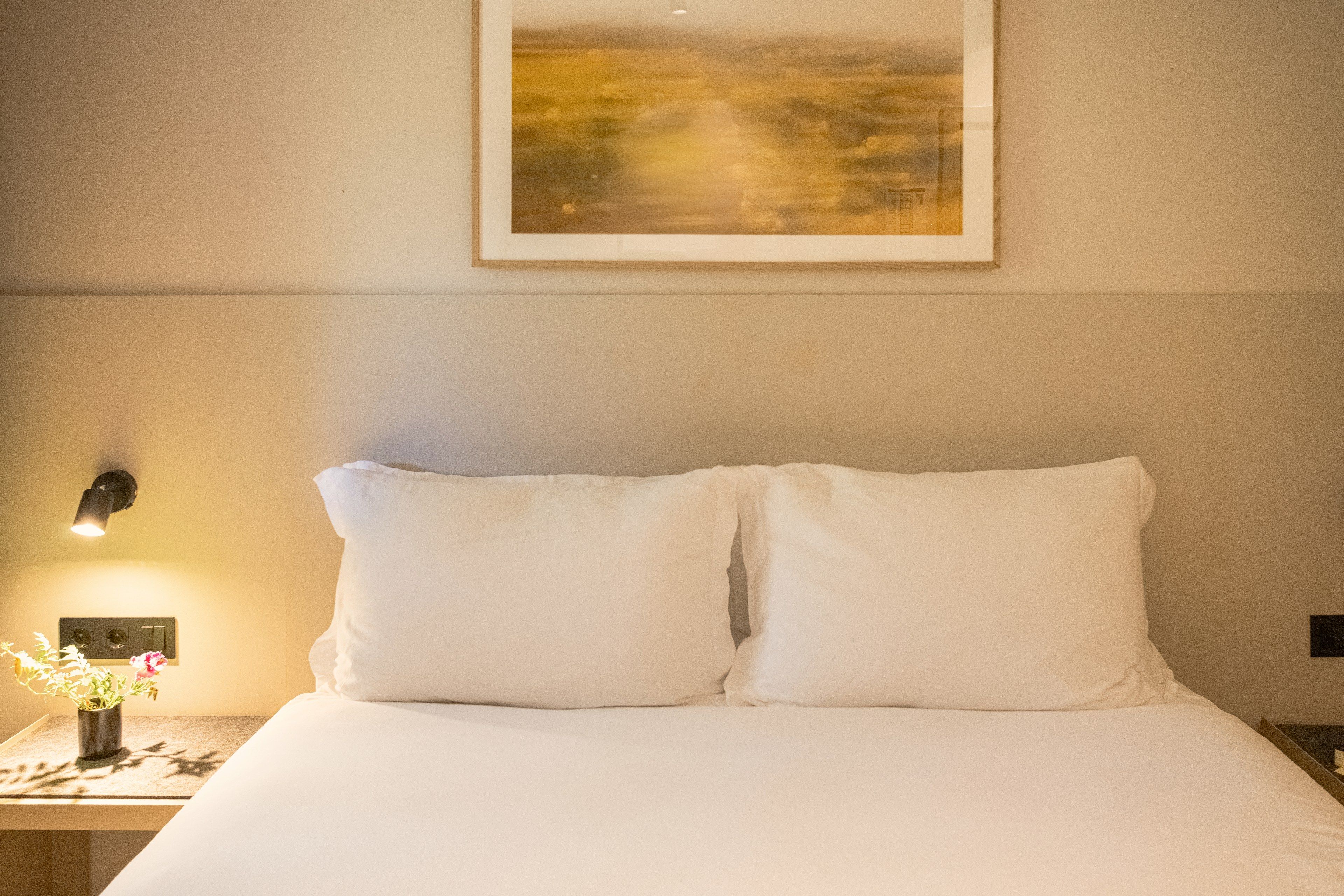 Habitació d'hotel amb llit, una tauleta de nit, una làmpada fixada a la paret i un quadre al mur.