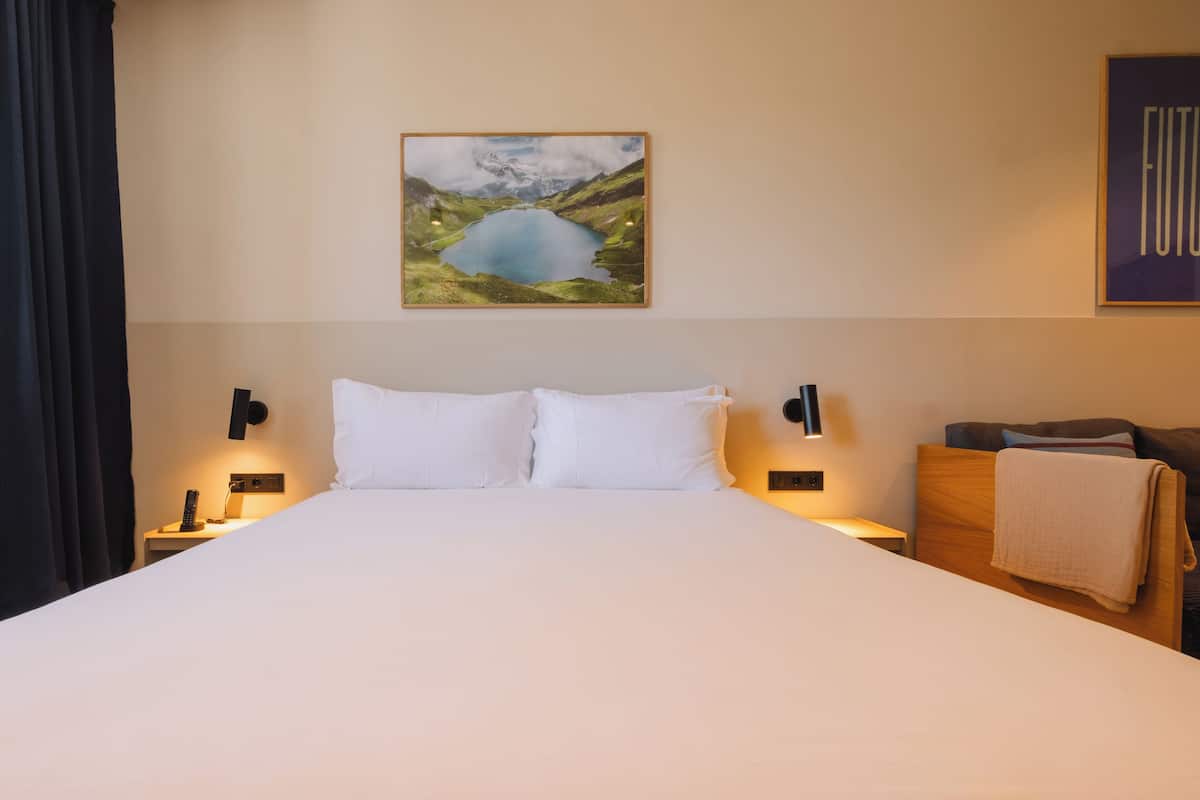 Habitació d'hotel amb un llit gran, dues taules de nit amb làmpades, un quadre d'un llac i una paraula emmarcada a la paret.