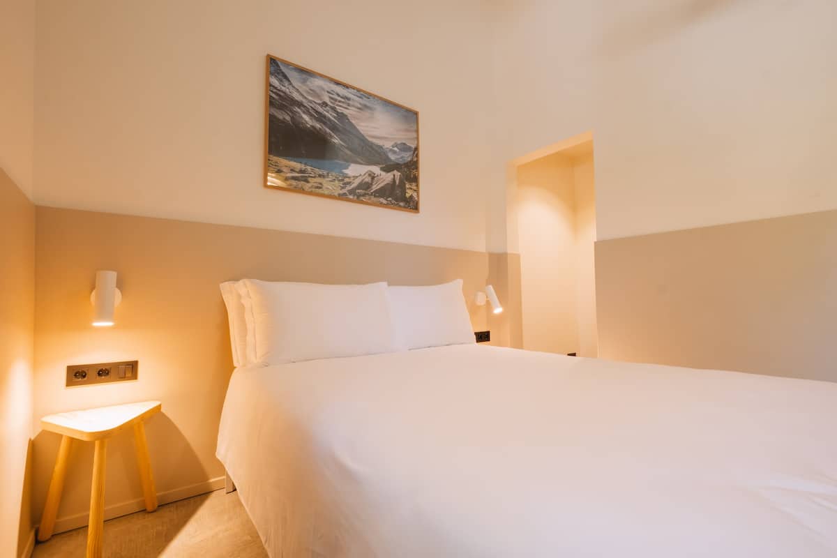 Llits ben fets a una habitació d'hotel, amb un quadre de paisatge de muntanya a sobre, una tauleta de nit amb una làmpada i una làmpada penjada a la paret.