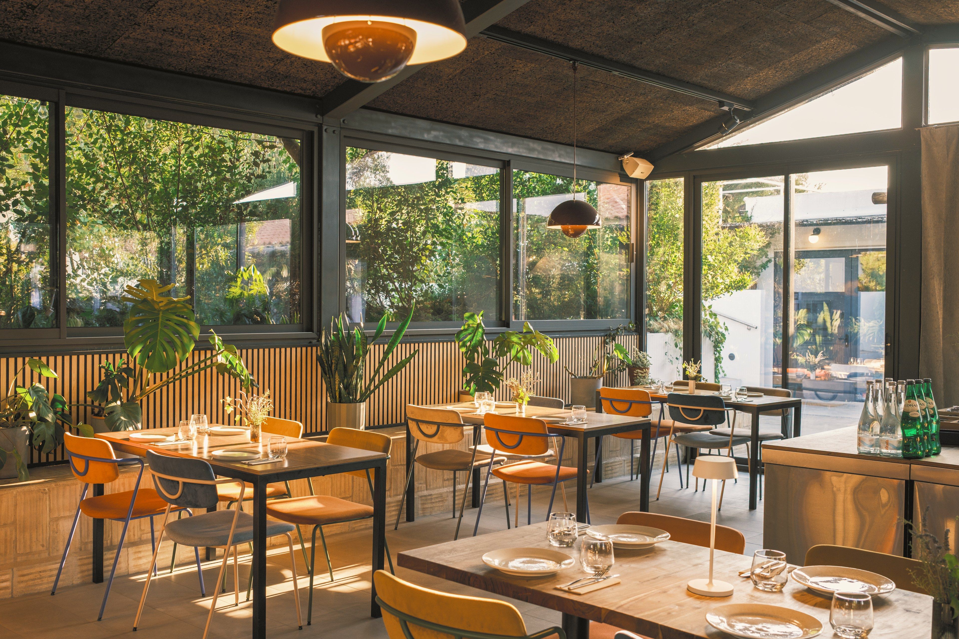 An indoor dining area with tables and chairs, large windows, and potted plants.