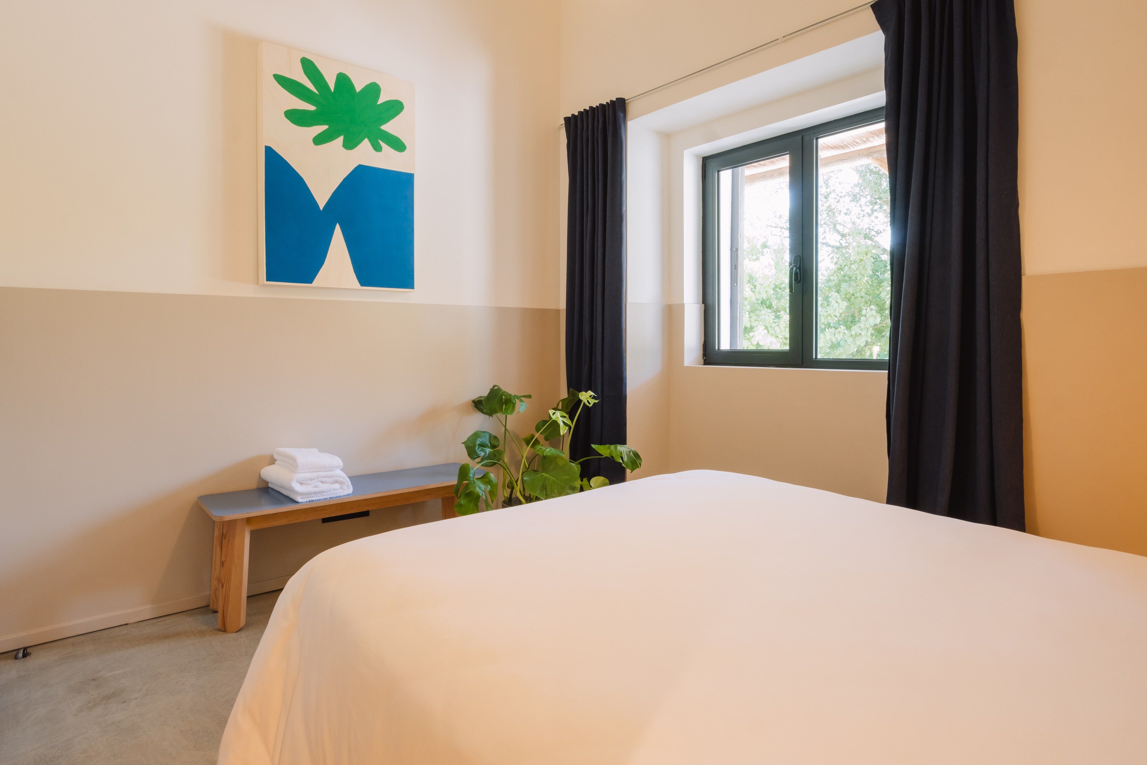 A hotel room with a bed, a bench, a potted plant, and a window with curtains.