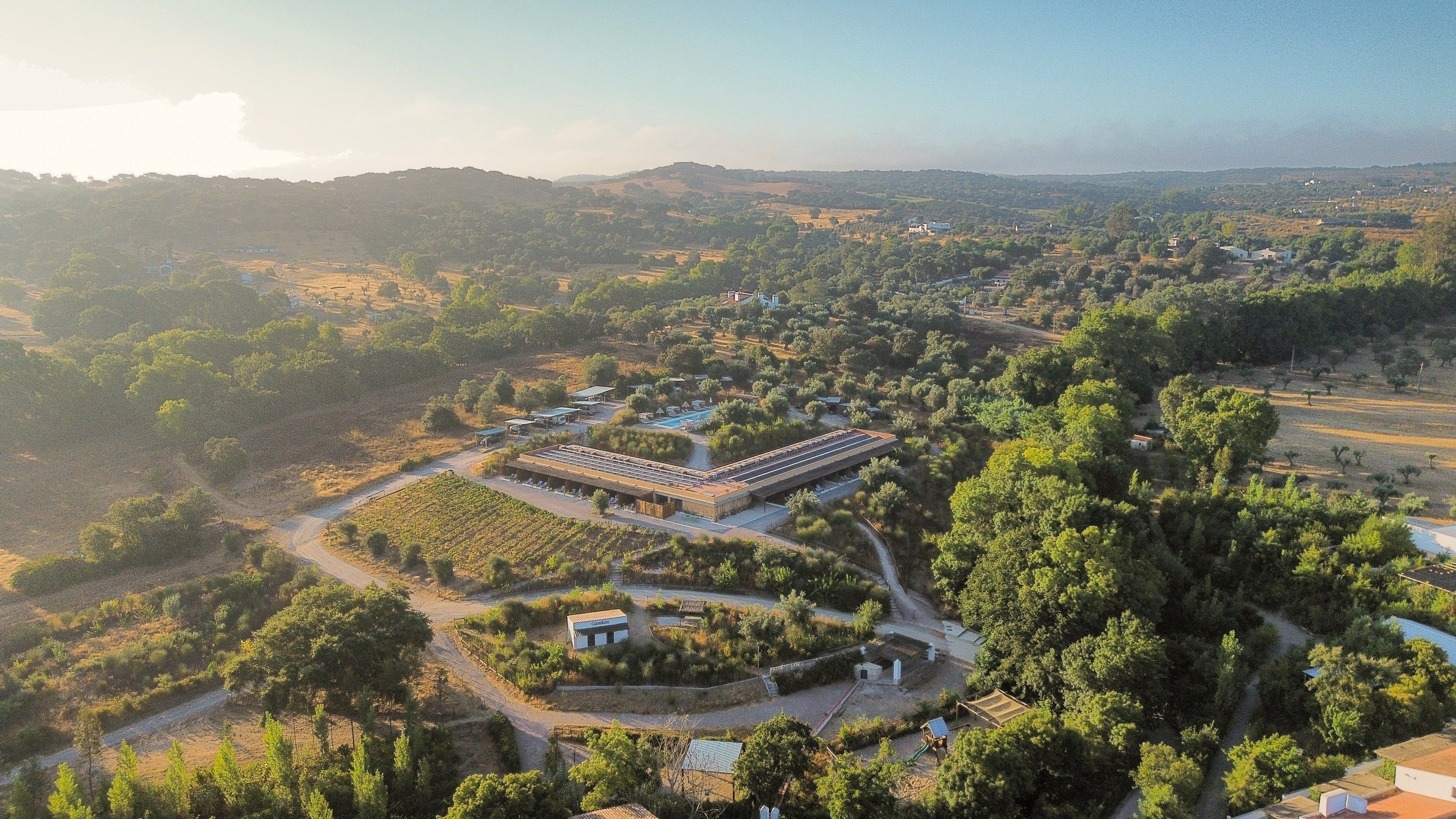 Vista aérea de uma propriedade com um edifício grande, jardins ao redor e uma paisagem montanhosa.