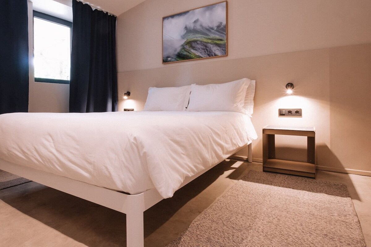 A neatly made bed with white linens in a modern room with a large window, a painting on the wall, and two bedside lamps.